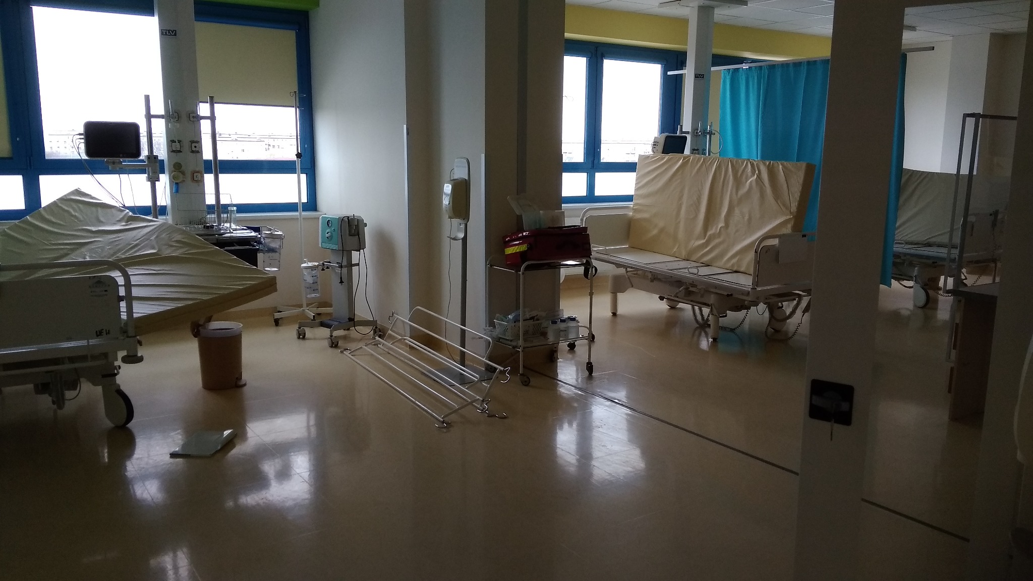 Pusta sala szpitalna z łóżkami, sprzętem medycznym, wózkiem z czerwonym pojemnikiem i metalowymi elementami na podłodze, widok z perspektywy wejścia.