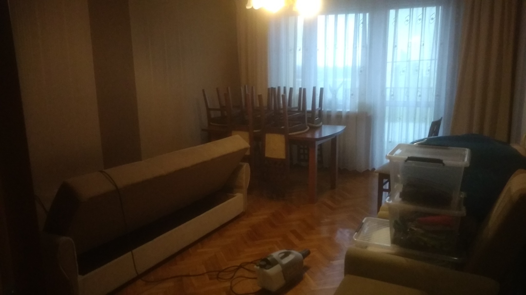 Pokój z meblami przygotowanymi do wyniesienia: odwrócone krzesła na stole, rozłożona sofa, plastikowe pojemniki spakowane na kanapie, odkurzacz na podłodze z parkietu w słabym oświetleniu.