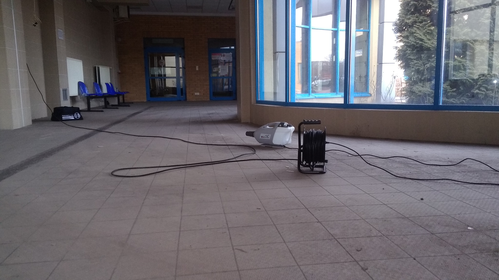 Zamgławiacz ULV i zwinięty kabel przed przeszkloną ścianą wewnątrz budynku, prawdopodobnie podczas dezynfekcji pomieszczenia.