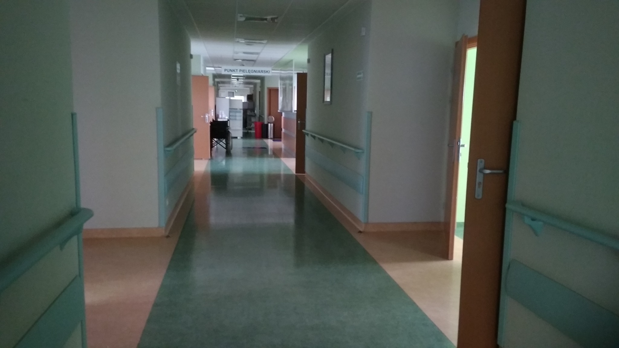 Długi, pusty korytarz szpitalny z zieloną podłogą i poręczami na ścianach, widok z perspektywy otwartych drzwi.