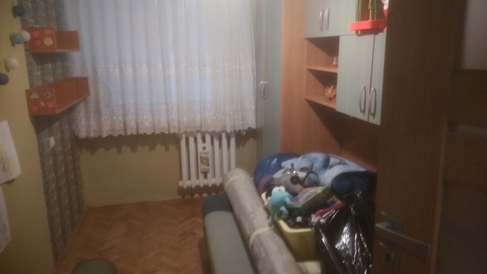 Pokój przed posprzątaniem: zwinięte dywany, zabawki i śmieci w koszu, widoczne meble i grzejnik pod oknem.