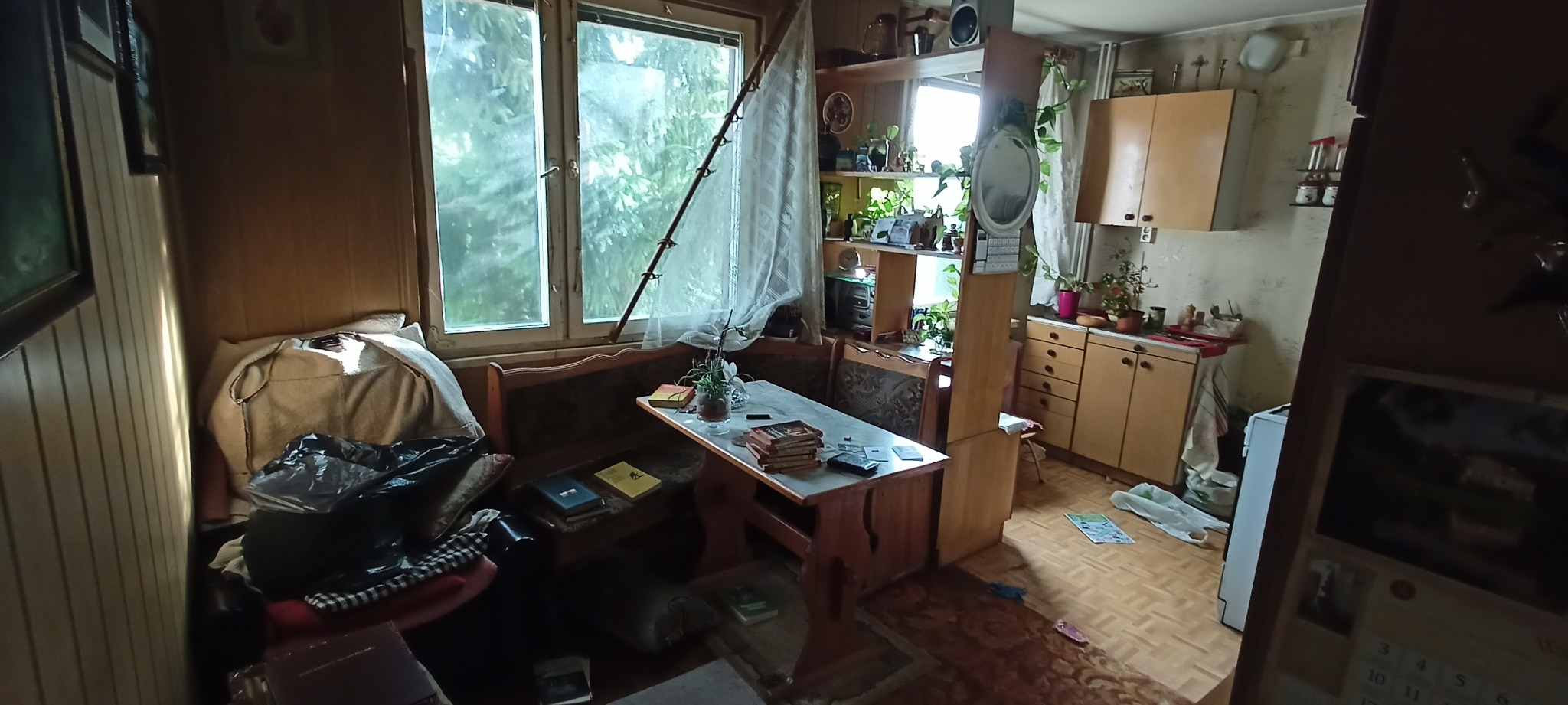 Widok na zagracone wnętrze mieszkania z drewnianymi meblami, oknem z firanką i roślinami doniczkowymi na półkach.