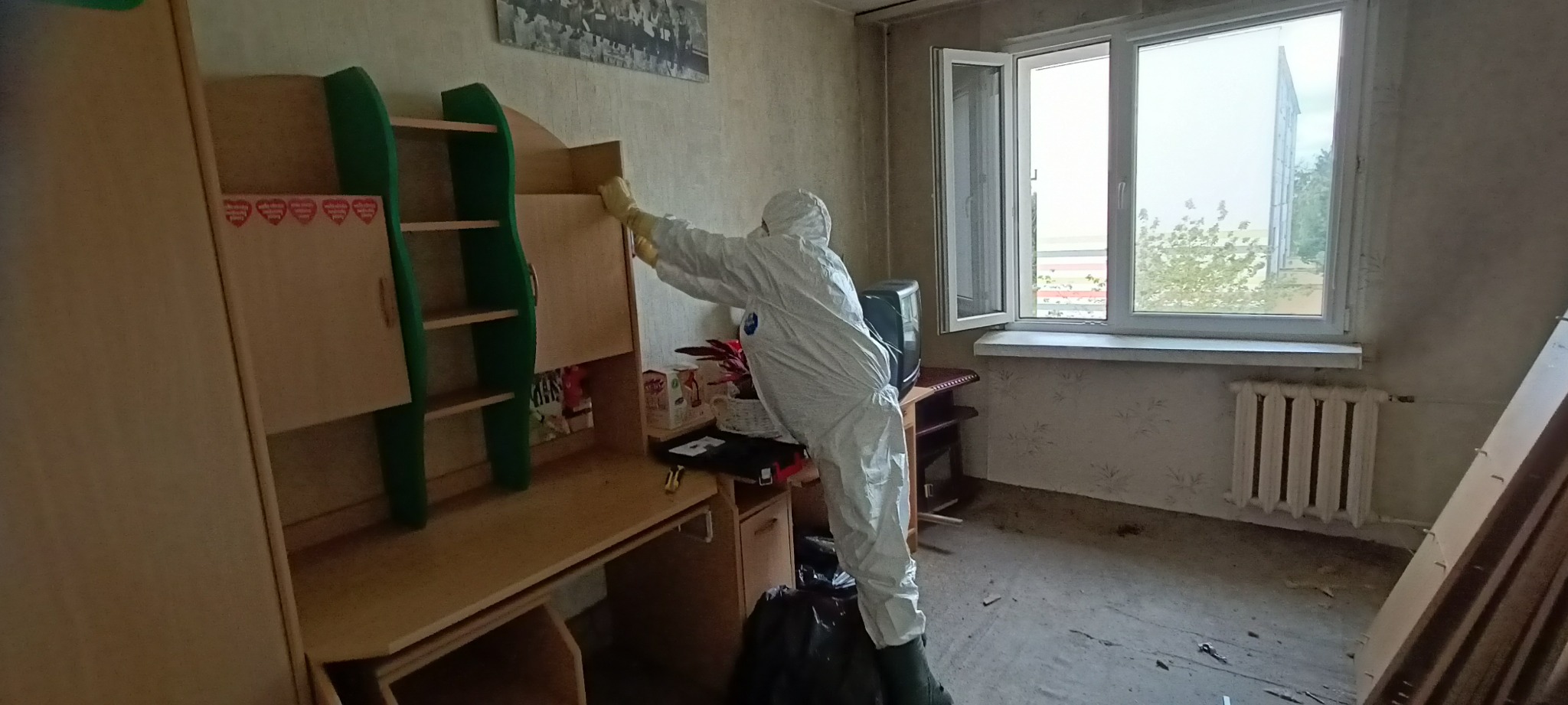 Osoba w kombinezonie ochronnym i rękawicach sprząta pokój dziecięcy, opróżniając półki regału z książkami i przedmiotami, widoczne ślady zabrudzeń na podłodze i ścianach.