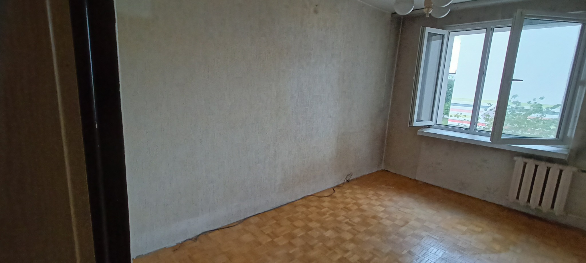 Pusty pokój z parkietem, otwartym oknem z widokiem na zieleń i grzejnikiem pod parapetem; ściany wymagają odświeżenia.