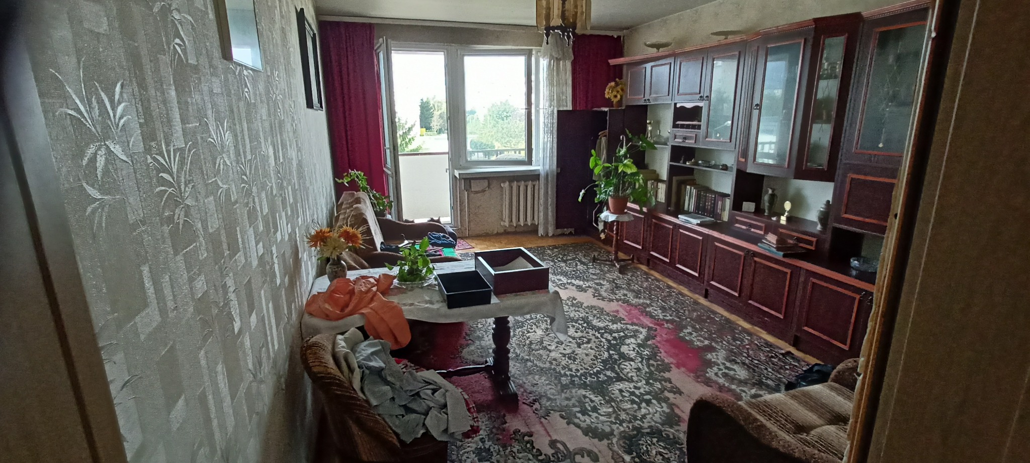 Pokój dzienny przed generalnym porządkiem: widoczny bałagan na stole, dywanie i krześle, na tle meblościanki i okna balkonowego.