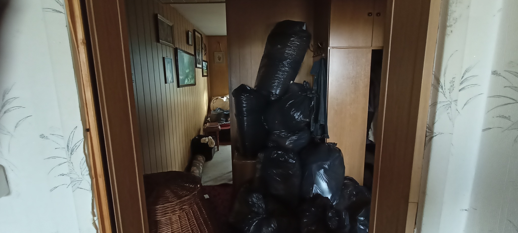 Stos czarnych worków na śmieci w otwartym wejściu do pokoju z boazerią i ramkami na ścianach, widoczne z korytarza z tapetą w roślinny wzór.