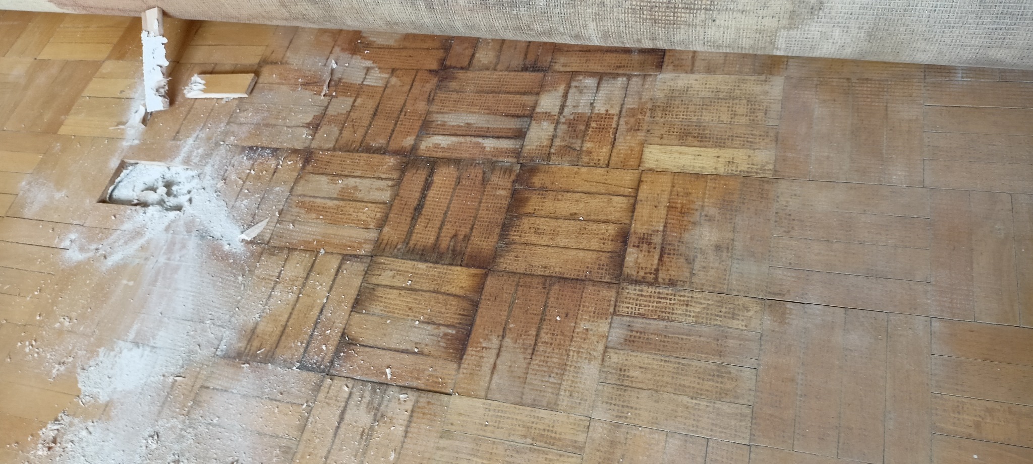 Fragment zniszczonej parkietowej podłogi w trakcie renowacji, z widocznym wyciętym fragmentem, resztkami kleju i warstwą pyłu po szlifowaniu.