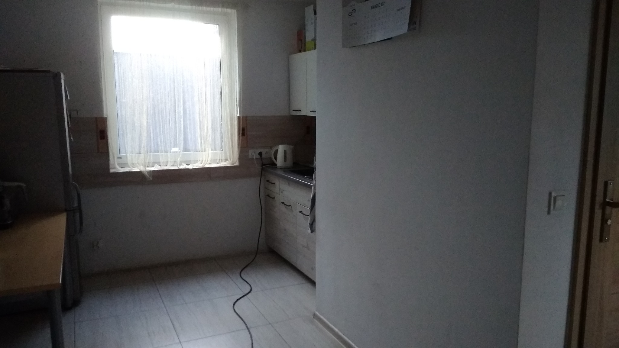 Kuchnia z oknem i firanką, widoczny fragment lodówki, białe szafki, czajnik elektryczny i kalendarz na ścianie.
