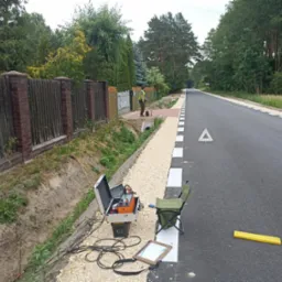 Prace geofizyczne na poboczu drogi: otwarty kufer z aparaturą pomiarową, zwinięte kable, krzesło turystyczne, żółty ogranicznik na asfalcie, w tle pracownik w kamizelce odblaskowej przy studzience...