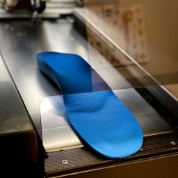 Gabinet Rehabilitacji i Ortopodologii mgr Jerzy Kowalski, mgr Robert Kowalski - Niebieska, trójwymiarowa wkładka ortopedyczna z widoczną teksturą, umieszczona wewnątrz urządzenia, prawdopodobnie drukarki 3D, z widocznymi elementami konstrukcyjnymi i oświetleniem studyjnym.