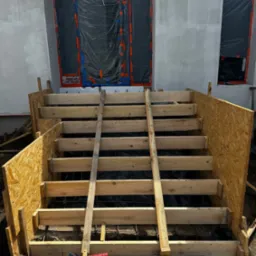 Konstrukcja drewnianego szalunku schodów zewnętrznych z widocznym zbrojeniem, przygotowana do wylania betonu przy budynku z oknami zabezpieczonymi folią.