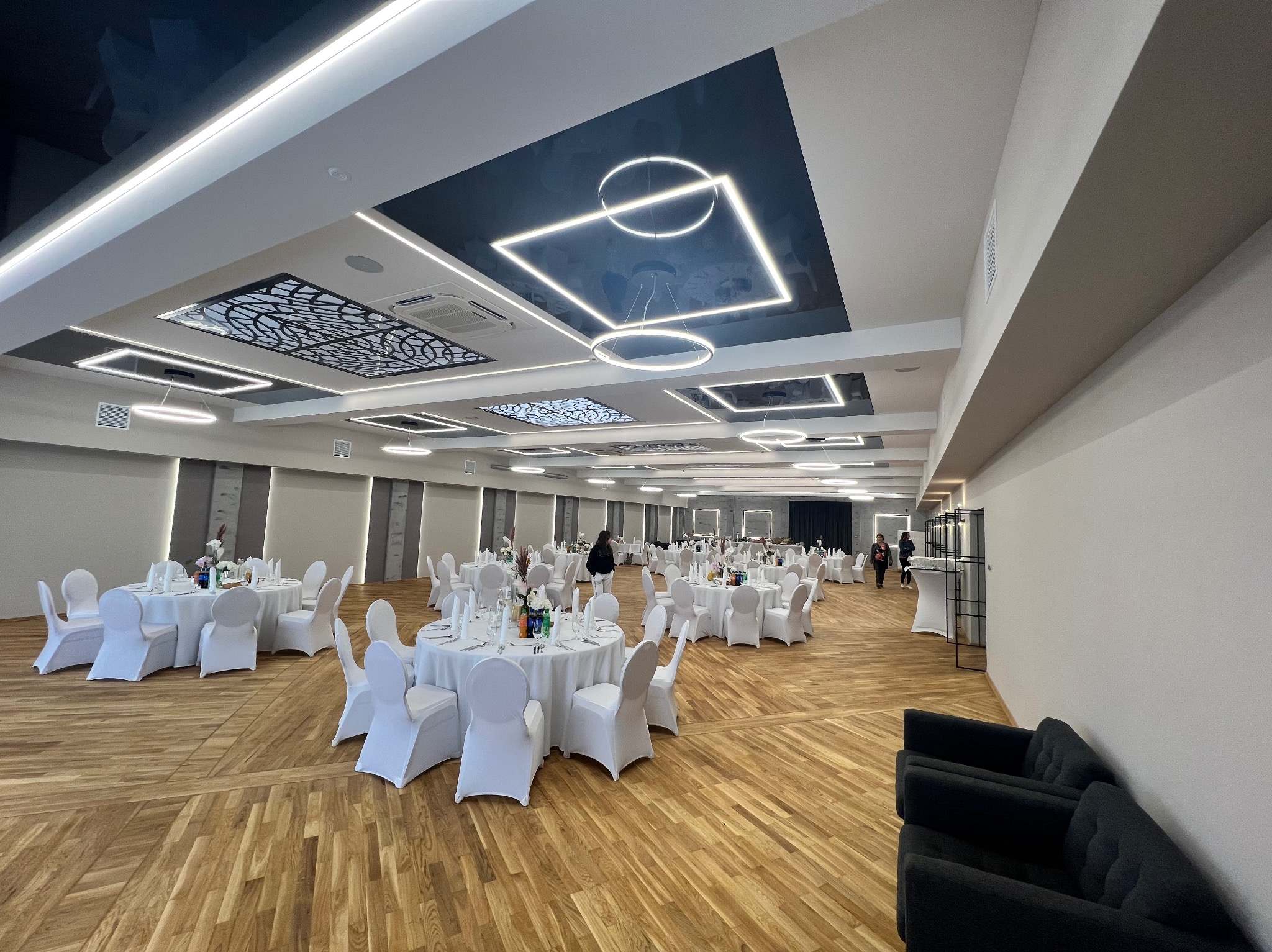Przestronna sala bankietowa z parkietem, białymi obrusami na okrągłych stołach i designerskim oświetleniem sufitowym z motywem geometrycznym.