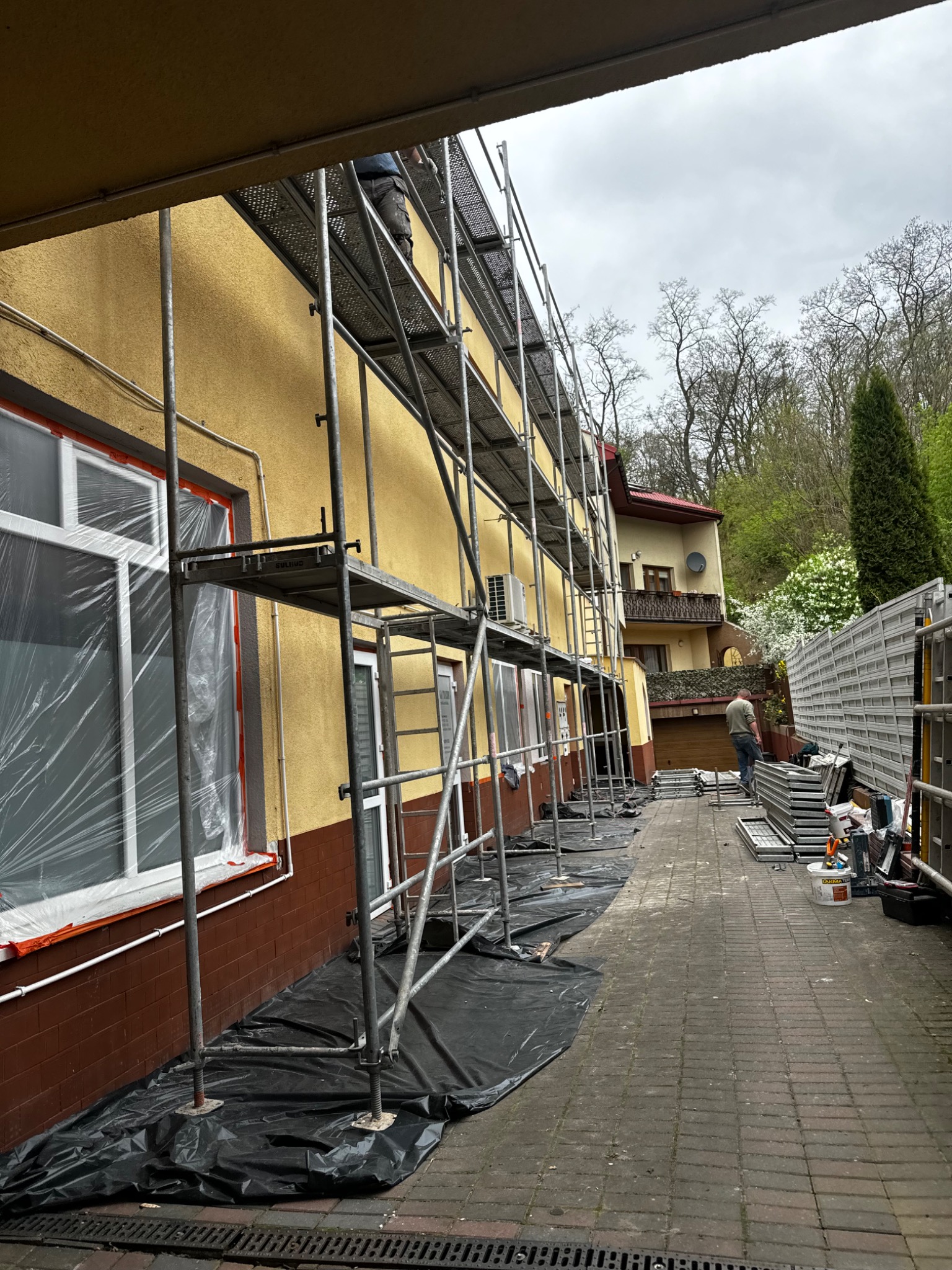 Dom jednorodzinny z żółtą elewacją w trakcie prac remontowych, z rozstawionym rusztowaniem i zabezpieczonymi oknami folią, widoczny fragment chodnika z narzędziami i materiałami budowlanymi.