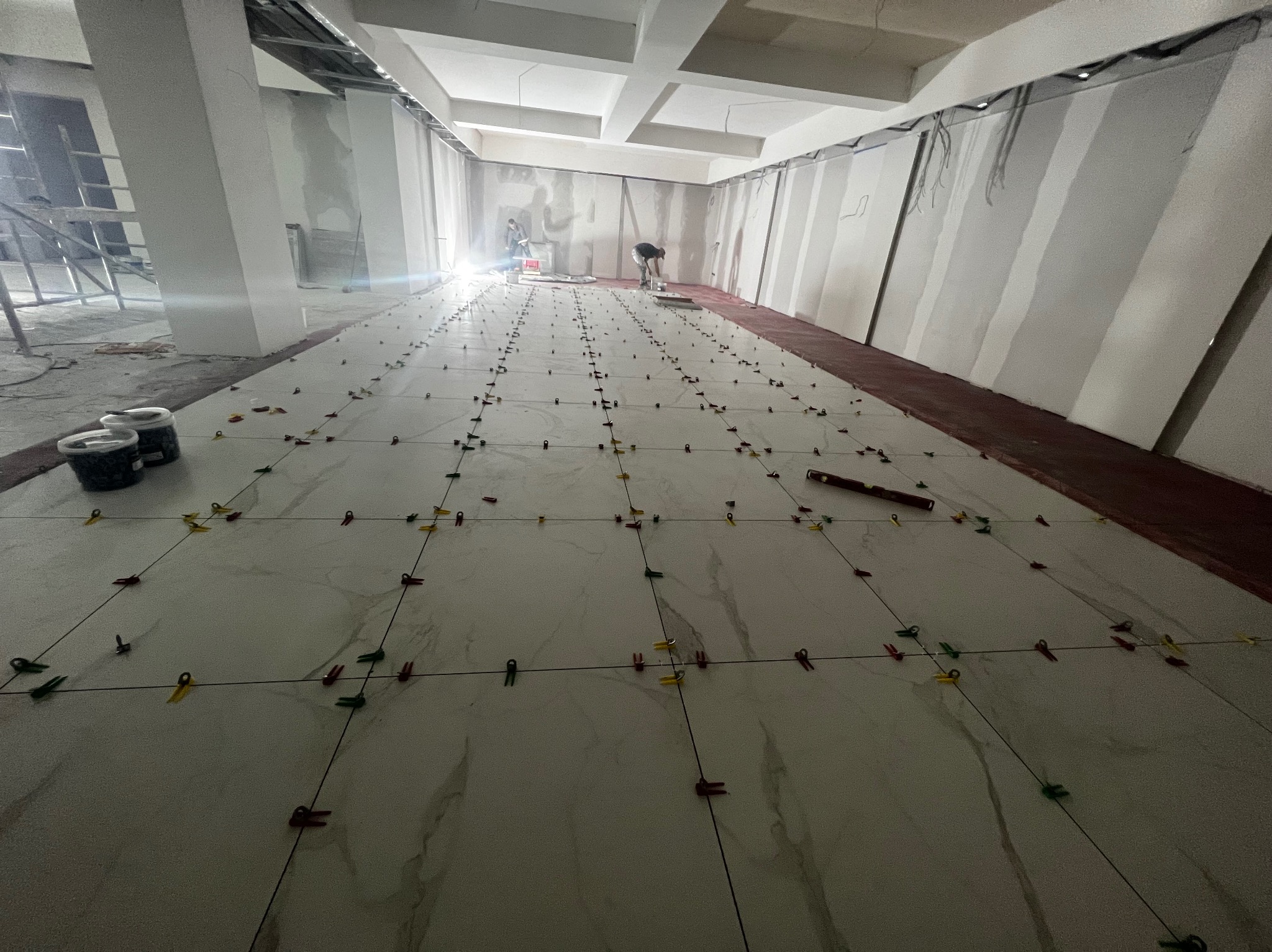 Układanie białych płytek podłogowych z marmurkowym wzorem w pomieszczeniu w trakcie wykańczania, widoczne kolorowe klipsy poziomujące, poziomica, puszki z farbą i pracownicy w tle.