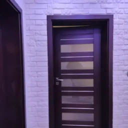 Nowoczesne drzwi wewnętrzne z poziomymi przeszkleniami, osadzone w ścianie wyłożonej dekoracyjną białą cegłą, oświetlone od góry subtelnym światłem LED.