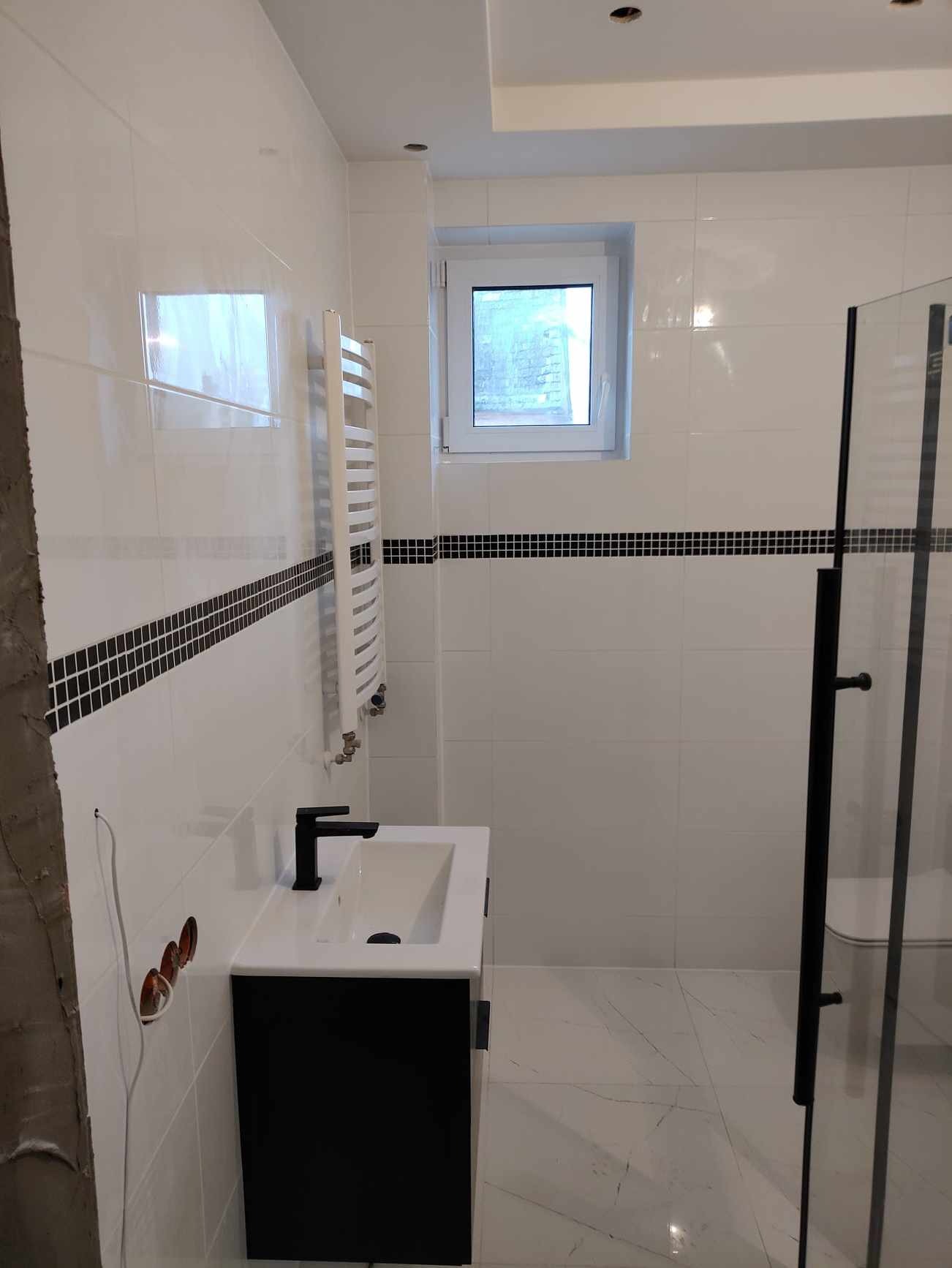 Wykończona łazienka z białymi płytkami, czarnym paskiem dekoracyjnym, czarną armaturą, grzejnikiem drabinkowym i kabiną prysznicową z czarnymi ramami, widoczne niedokończone prace przy instalacji...