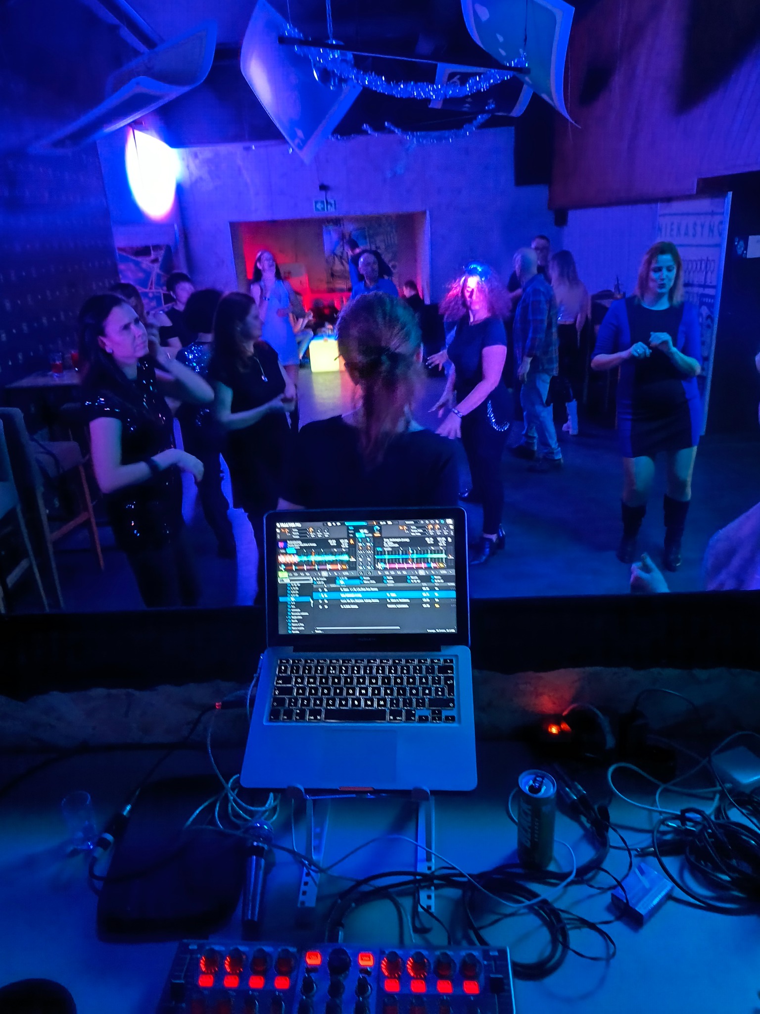 DJ z laptopem i kontrolerem na stanowisku, w tle tańczący tłum w niebieskim oświetleniu klubu.