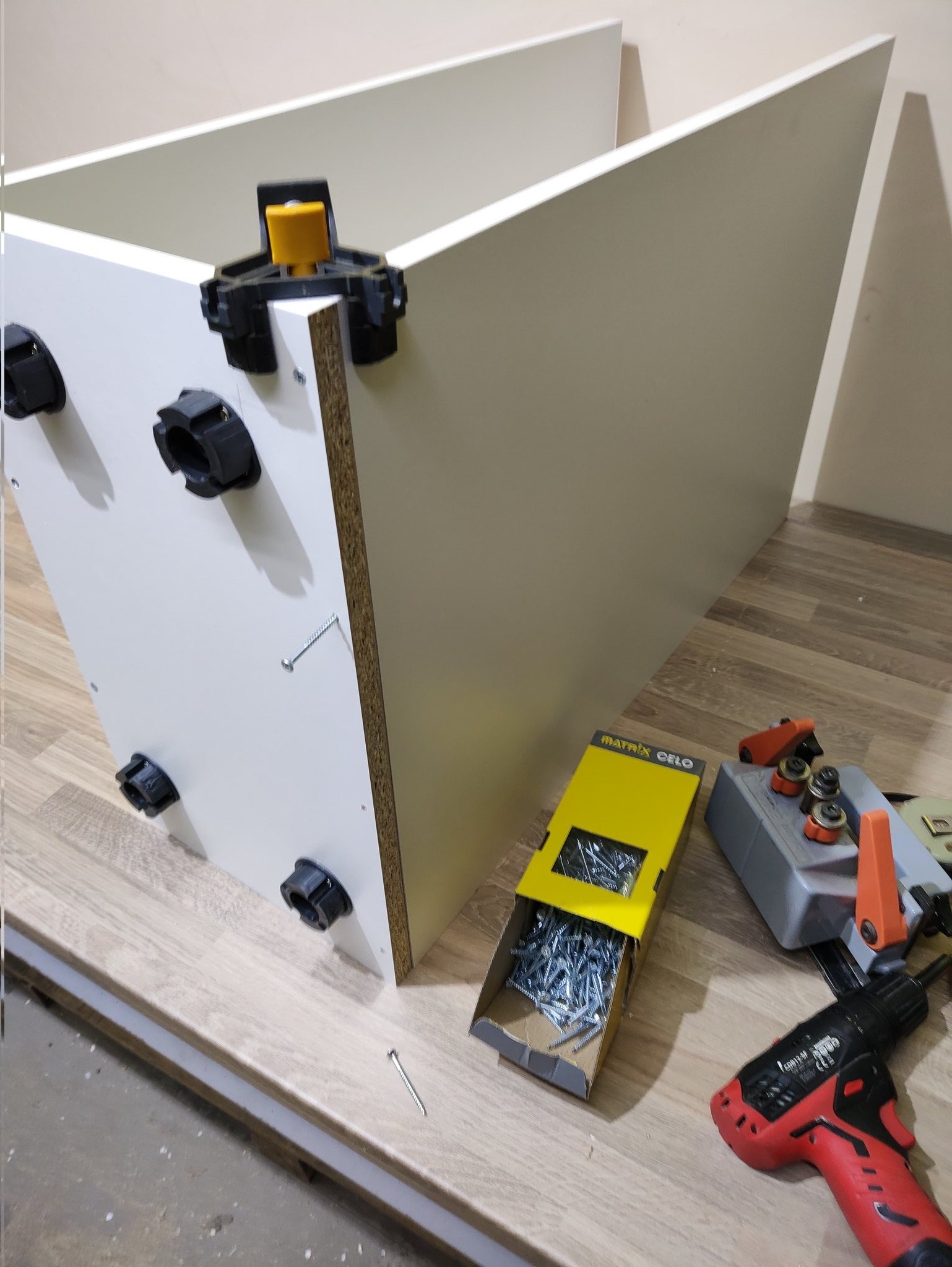 Fragment montażu białej szafy z widocznymi elementami mocującymi, wkrętami w otwartym pudełku i wkrętarką na podłodze.