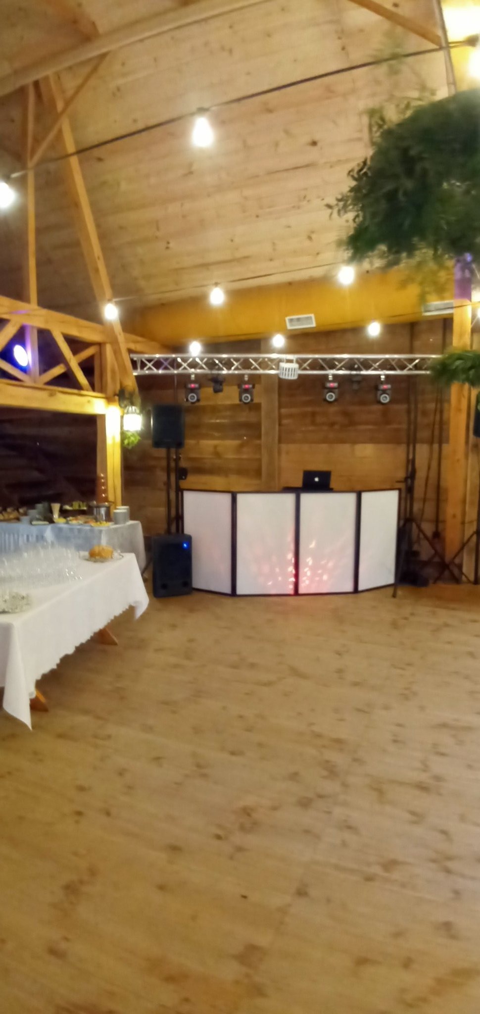 Scena z nagłośnieniem i oświetleniem przygotowana na imprezę w drewnianej sali z cateringiem, widoczne głośniki, reflektory i parawan DJ-a