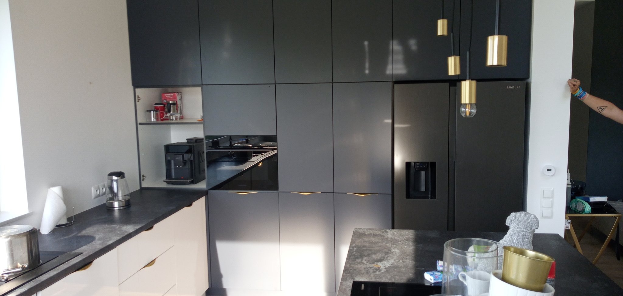 Nowoczesna kuchnia z szarymi, matowymi frontami szafek, złotymi uchwytami i wiszącymi lampami z elementami w kolorze złota. Widoczny ekspres do kawy, czajnik elektryczny i lodówka Samsung.