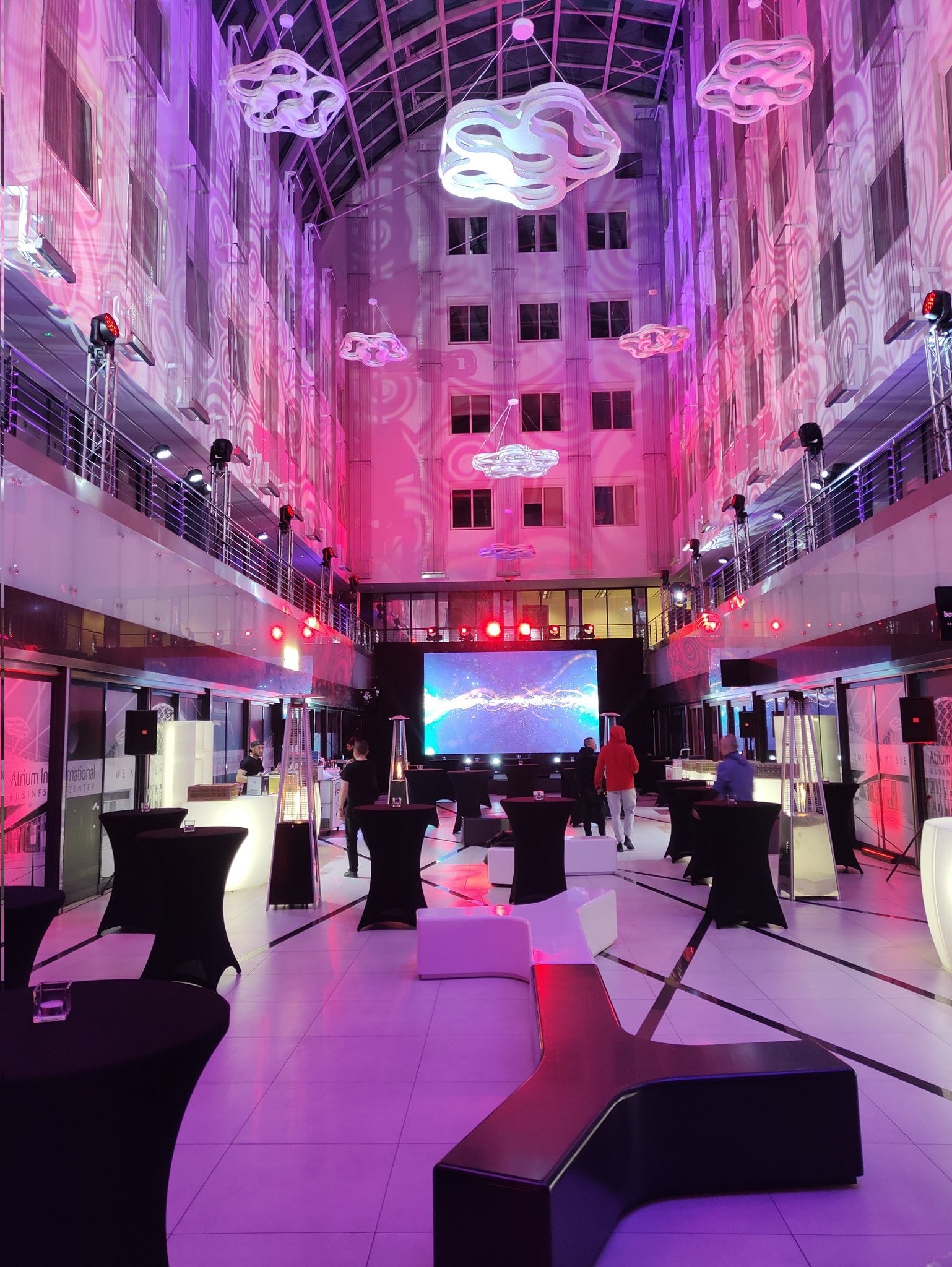 Wnętrze atrium oświetlone dekoracyjnym światłem w kolorach różu i fioletu, z podwieszonymi lampami w kształcie chmur, nagłośnieniem na balustradach i stolikami koktajlowymi przygotowanymi...