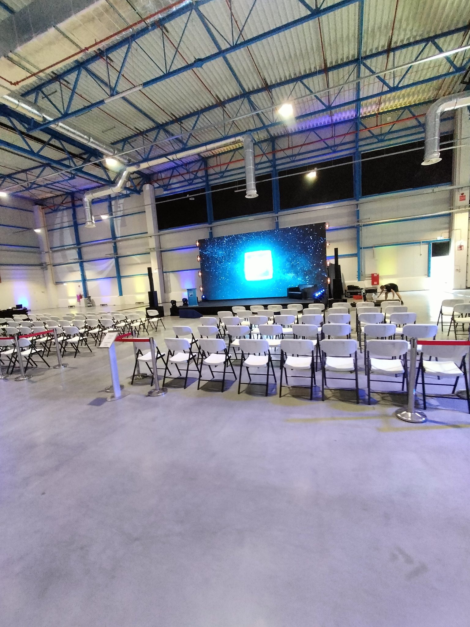 Sala konferencyjna z rzędami krzeseł ustawionych przed sceną z dużym ekranem wyświetlającym animację kosmosu, widoczne elementy konstrukcji dachu.