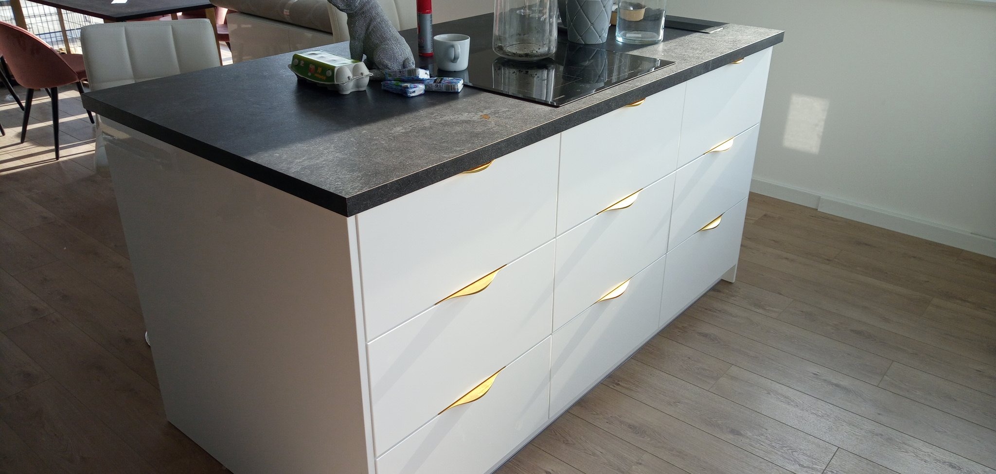 Wyspa kuchenna z białymi frontami szuflad z nietypowymi złotymi uchwytami, ciemny blat, płyta indukcyjna i akcesoria kuchenne.