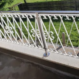 Biała, ozdobna balustrada balkonowa z motywami roślinnymi, wsparta na nierdzewnych słupkach, widok na zielony trawnik i drewniane ogrodzenie w tle.