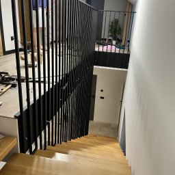 Dan-Technnik Daniel Hoszek - Montaż czarnej, pionowej balustrady na schodach z jasnego drewna; widoczne narzędzia i osoba w tle.