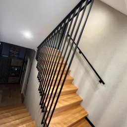 Dan-Technnik Daniel Hoszek - Nowoczesne schody drewniane z czarną, minimalistyczną balustradą metalową. Ujęcie z dołu, podkreślające geometrię i kontrast materiałów.