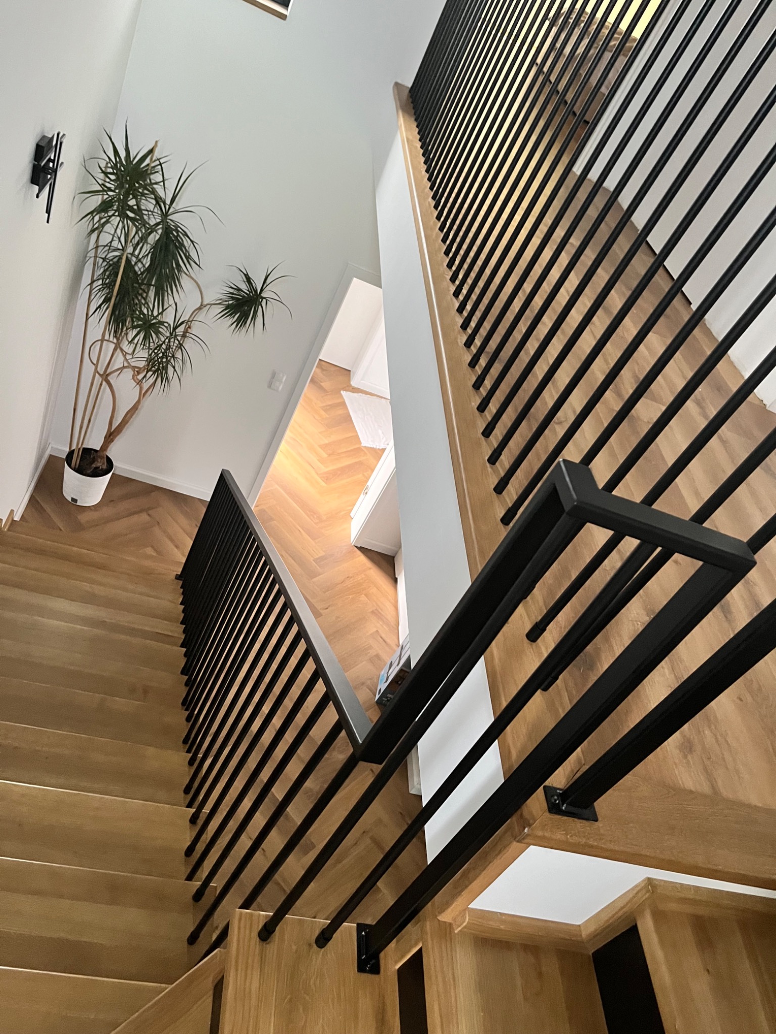 Nowoczesne schody drewniane z czarną, minimalistyczną balustradą z prętów, widok z góry na parkiet w jodełkę i roślinę doniczkową przy ścianie.