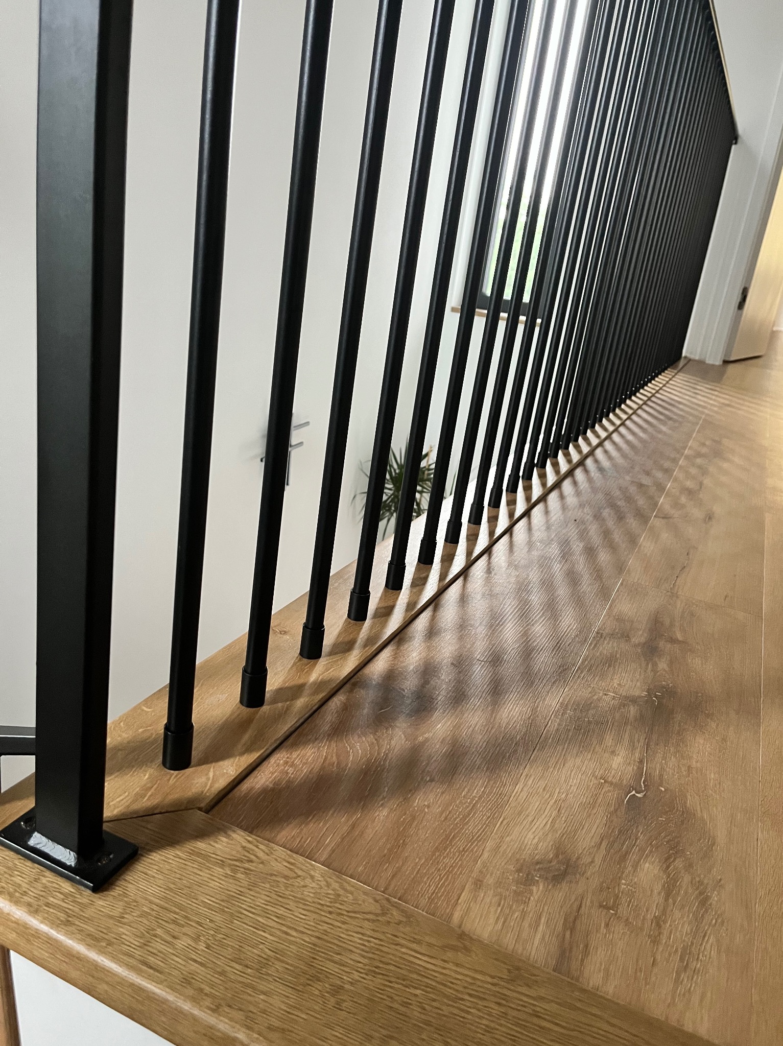 Nowoczesna balustrada schodowa z czarnymi, pionowymi słupkami i drewnianym wykończeniem, widok z bliska na połączenie słupków z podłogą, jasne światło podkreśla strukturę drewna.