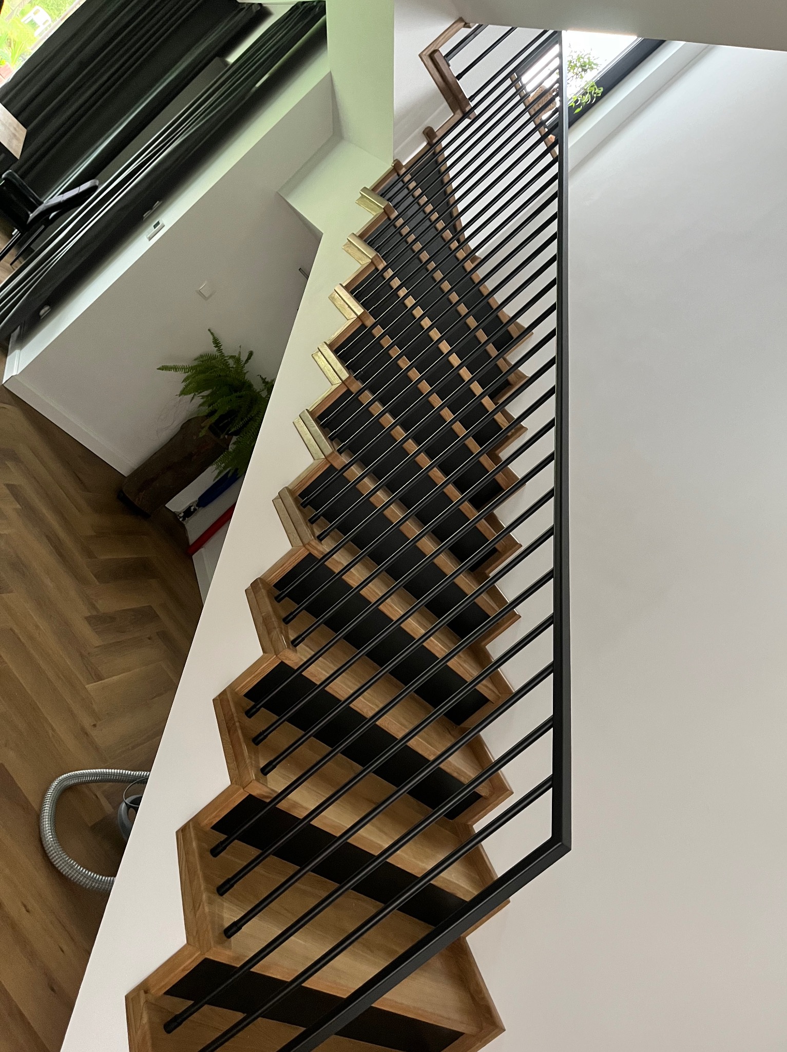 Nowoczesne schody z drewnianymi stopniami i czarnymi metalowymi barierkami, widok z góry, jasne wnętrze z parkietem w jodełkę i rośliną doniczkową w tle.