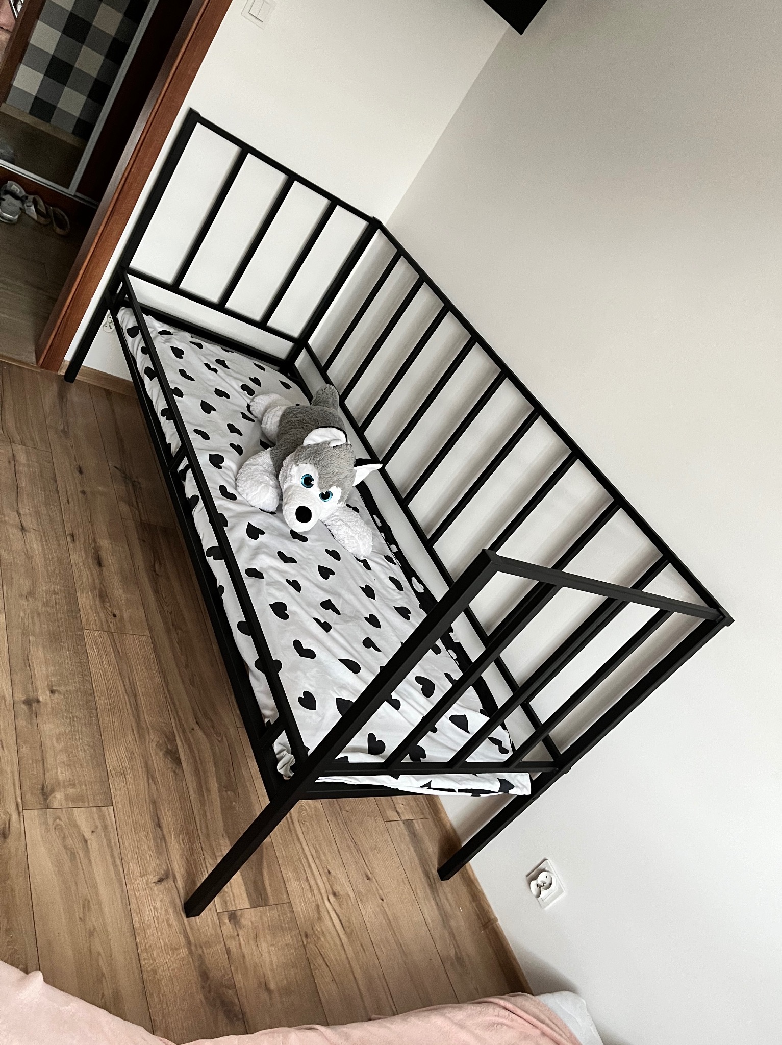 Minimalistyczne łóżko metalowe z czarną ramą, pościel w czarne serca i pluszowy piesek w pokoju z drewnianą podłogą.