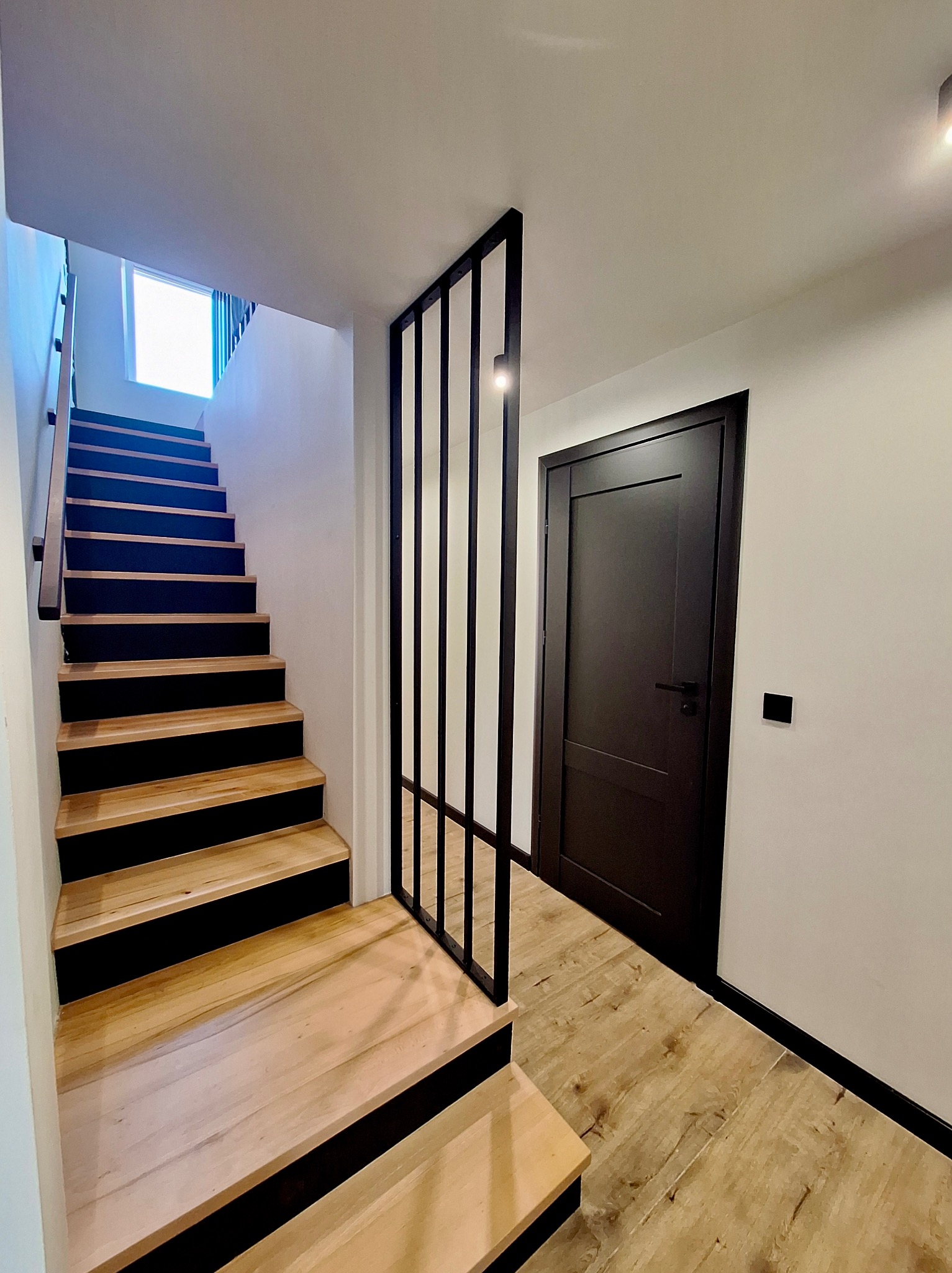 Wnętrze domu z drewnianymi schodami z czarnymi podstopnicami, czarną, minimalistyczną ścianką działową z metalowych profili oraz czarnymi drzwiami, oświetlone światłem dziennym i lampą sufitową.