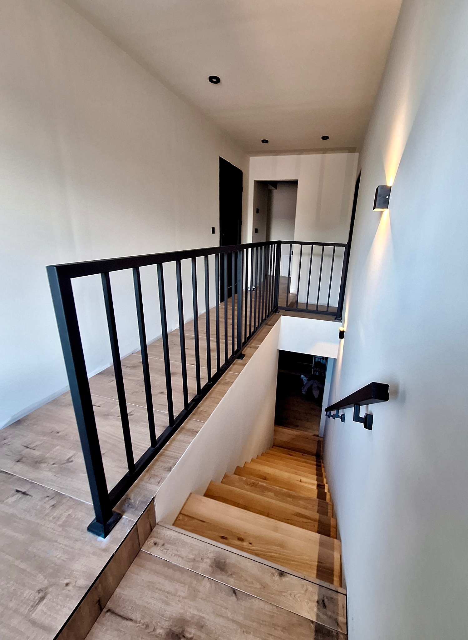 Nowoczesne wnętrze z drewnianymi schodami i czarną, minimalistyczną balustradą metalową oraz poręczą, oświetlone kinkietami ściennymi.