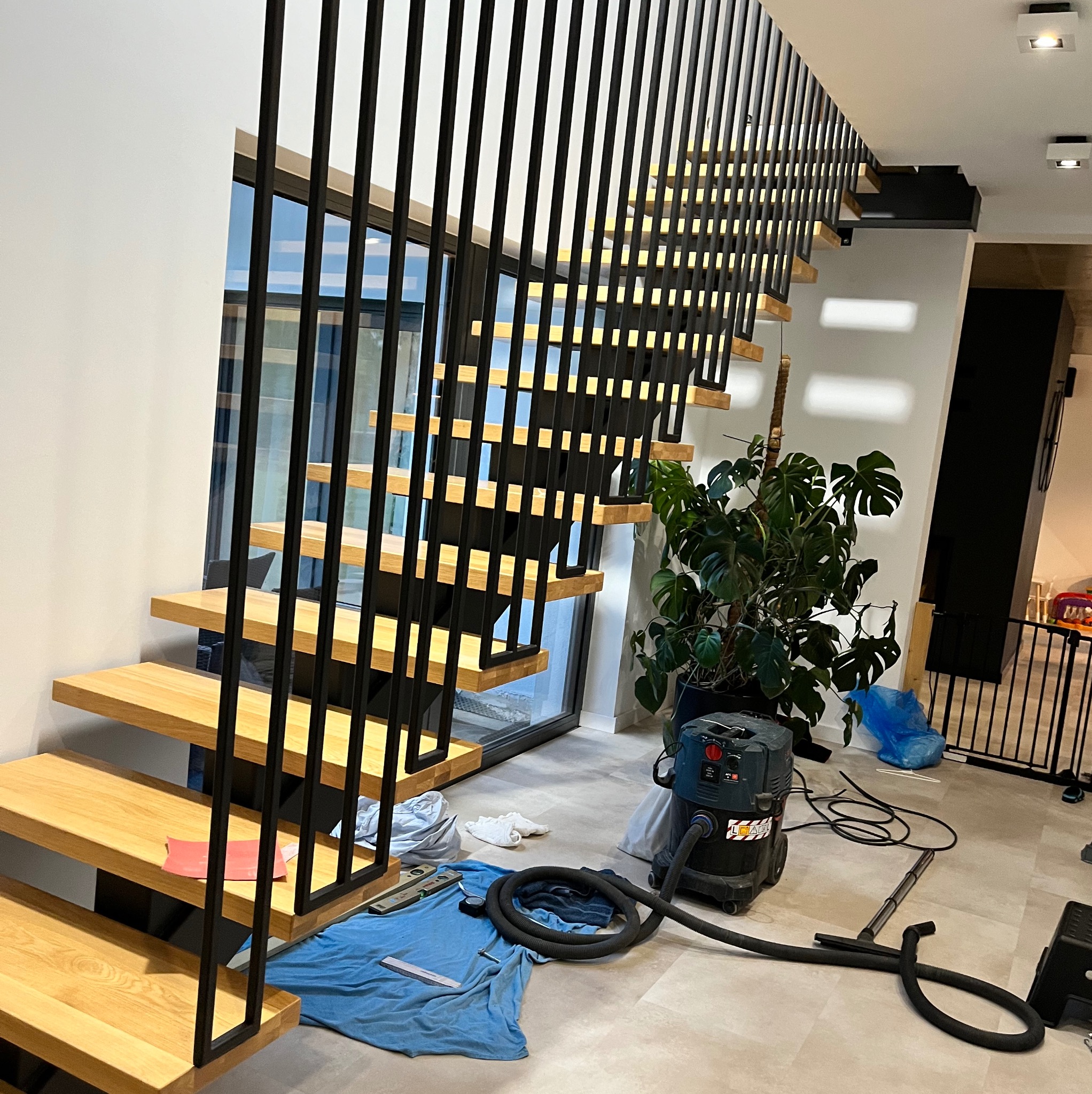 Nowoczesne schody drewniane z czarnymi, pionowymi balustradami, widoczne narzędzia i akcesoria czyszczące po montażu, duża roślina doniczkowa w tle.