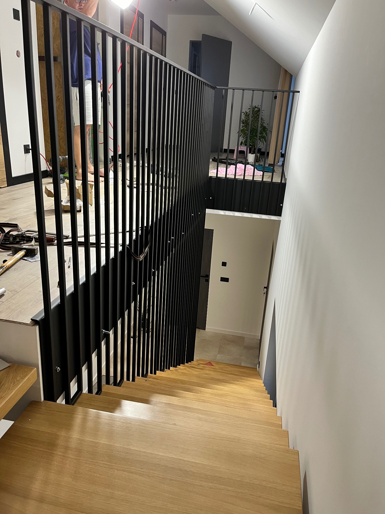 Montaż czarnej, pionowej balustrady na schodach z jasnego drewna; widoczne narzędzia i osoba w tle.