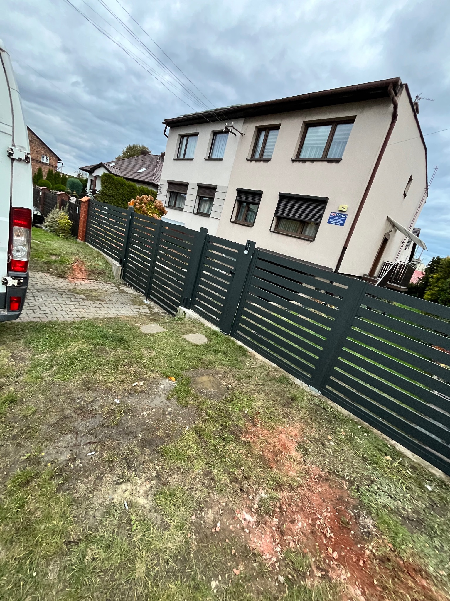 Nowoczesne, poziome ogrodzenie panelowe w kolorze grafitowym, oddzielające posesję od chodnika. Widoczny fragment białego busa i dwupiętrowy dom w tle.