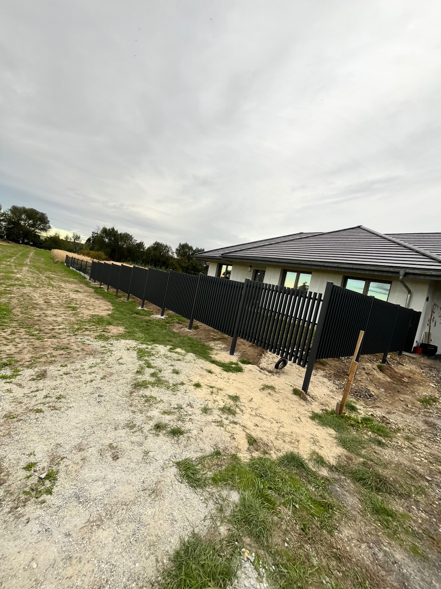 Ciemne, pionowe panele ogrodzeniowe z metalu, otaczające posesję z domem o szarym dachu. Widok z boku, z polną drogą i zielenią w tle, pochmurne niebo.