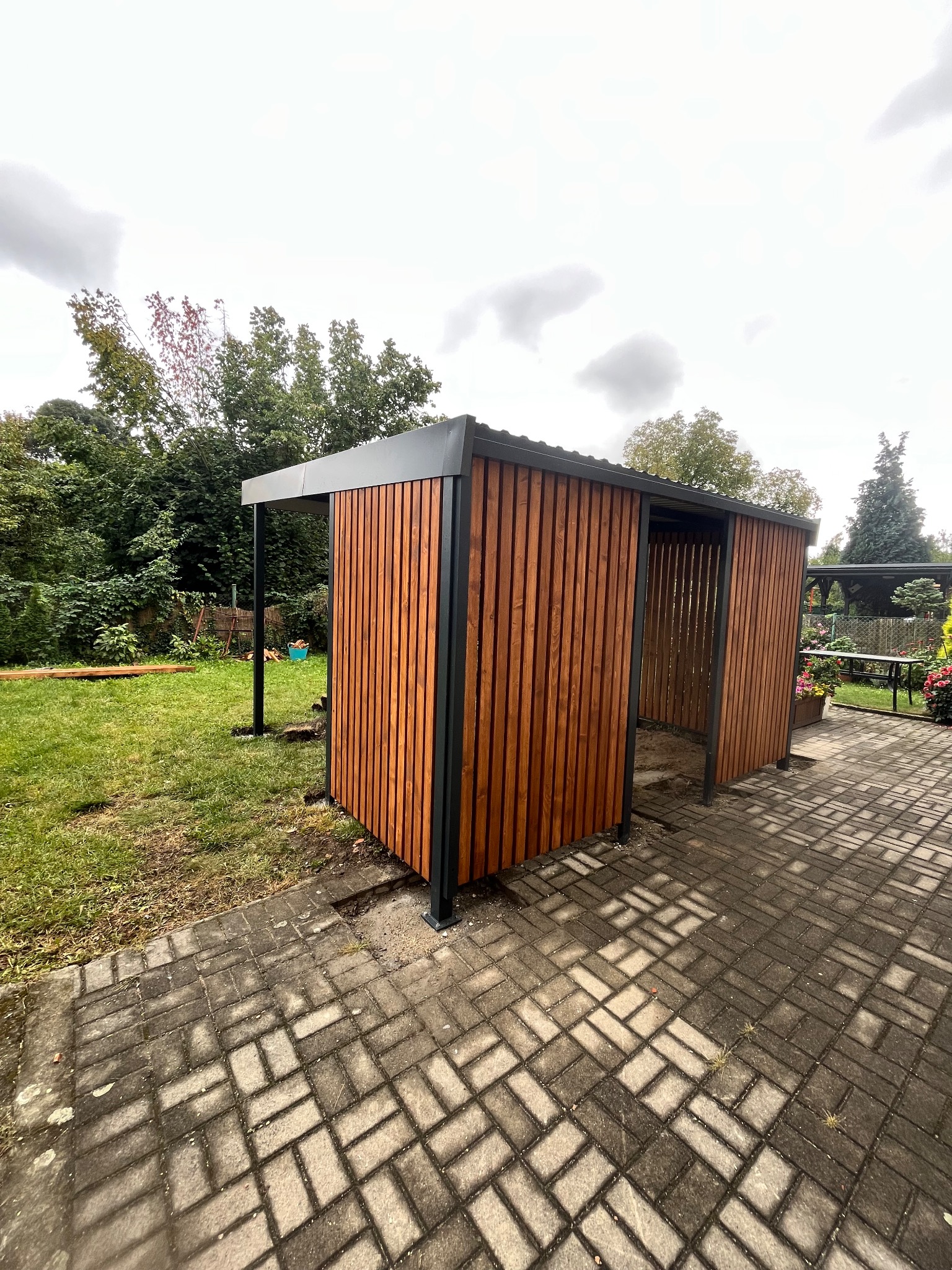 Nowoczesna pergola metalowa z drewnianym wypełnieniem, widok z zewnątrz na tle zielonego ogrodu i kostki brukowej. Konstrukcja w kolorze grafitowym, drewno w ciepłym odcieniu brązu.