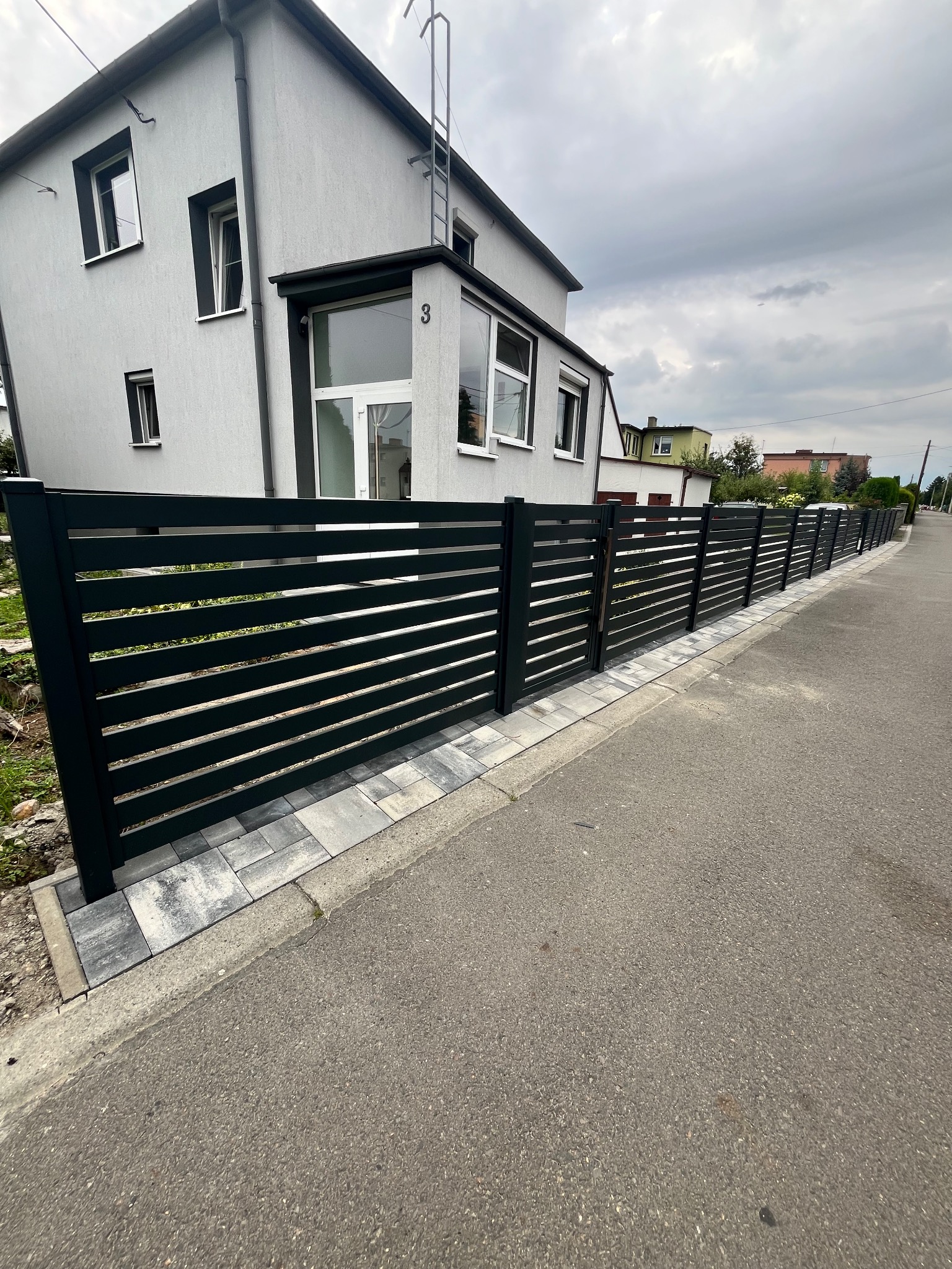 Nowoczesne, poziome ogrodzenie metalowe w kolorze antracytowym, z szarym brukiem u podstawy, na tle domu z jasną elewacją i pochmurnego nieba.
