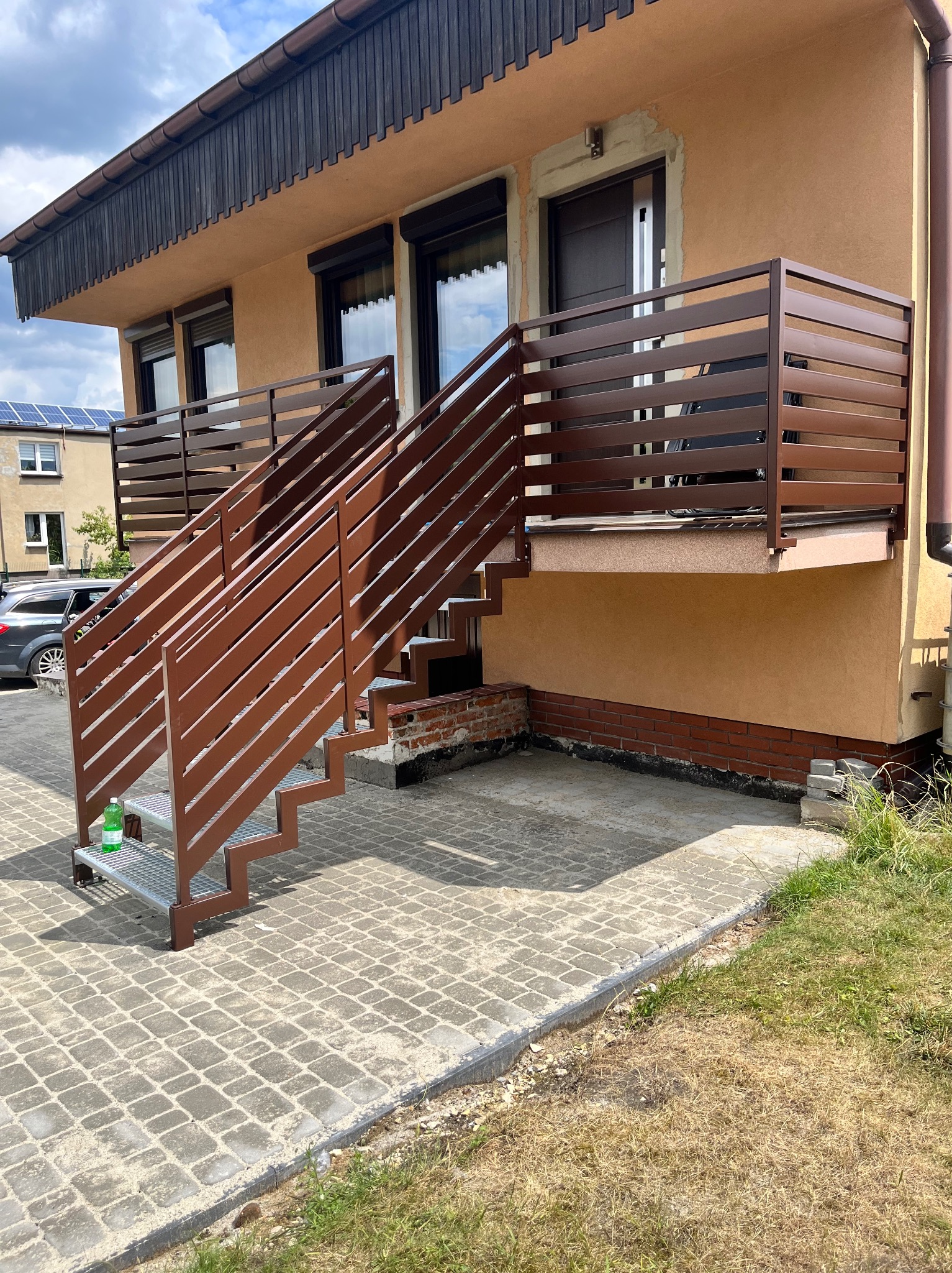 Brązowe schody metalowe z balustradą prowadzące na balkon domu. Widoczne poziome elementy balustrady i metalowe stopnie. Na pierwszym planie butelka wody.
