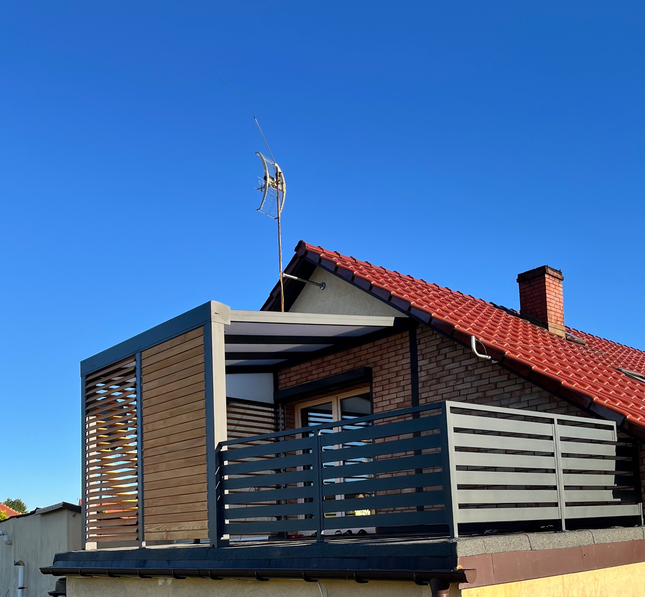 Nowoczesna pergola z drewnianymi żaluzjami i metalową balustradą na dachu budynku z czerwoną dachówką, na tle błękitnego nieba.