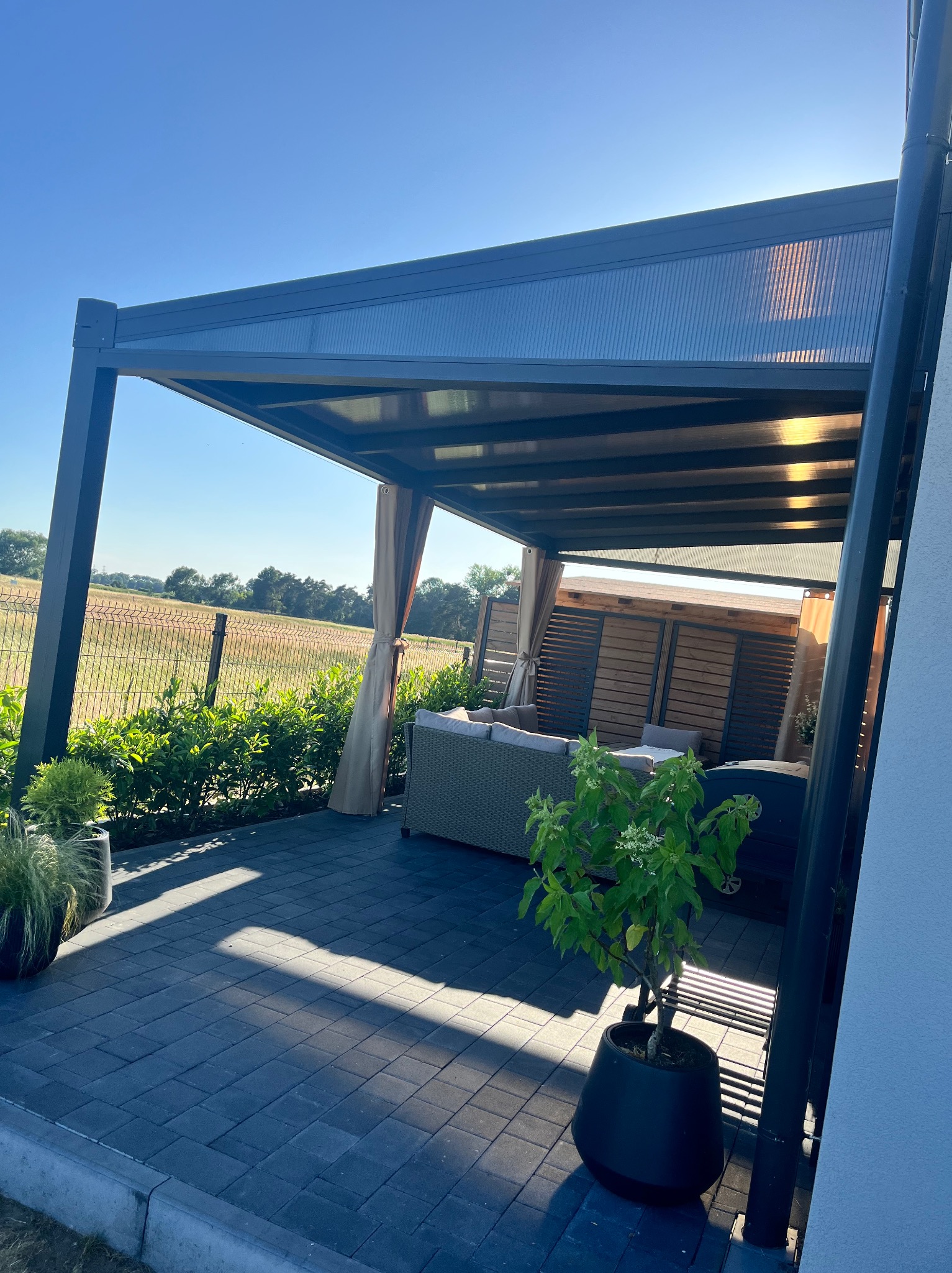 Nowoczesna pergola metalowa z zasłonami, oświetlona promieniami słońca, tworząca zacienioną strefę relaksu na tarasie z widokiem na zielone pola.