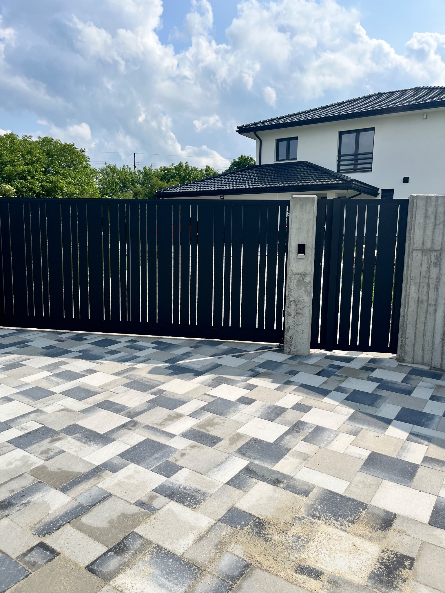 Nowoczesna, czarna brama wjazdowa z pionowymi szczeblami, osadzona na betonowych słupkach. Kostka brukowa w odcieniach szarości i bieli prowadzi do domu z ciemnym dachem.