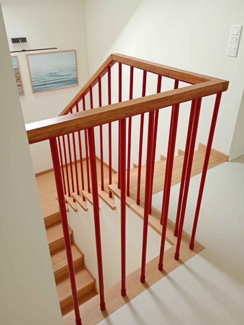 Nowoczesne schody z drewnianymi stopniami i czerwoną balustradą z drewnianym poręczą, widziane z góry, z obrazem morza na ścianie w tle.