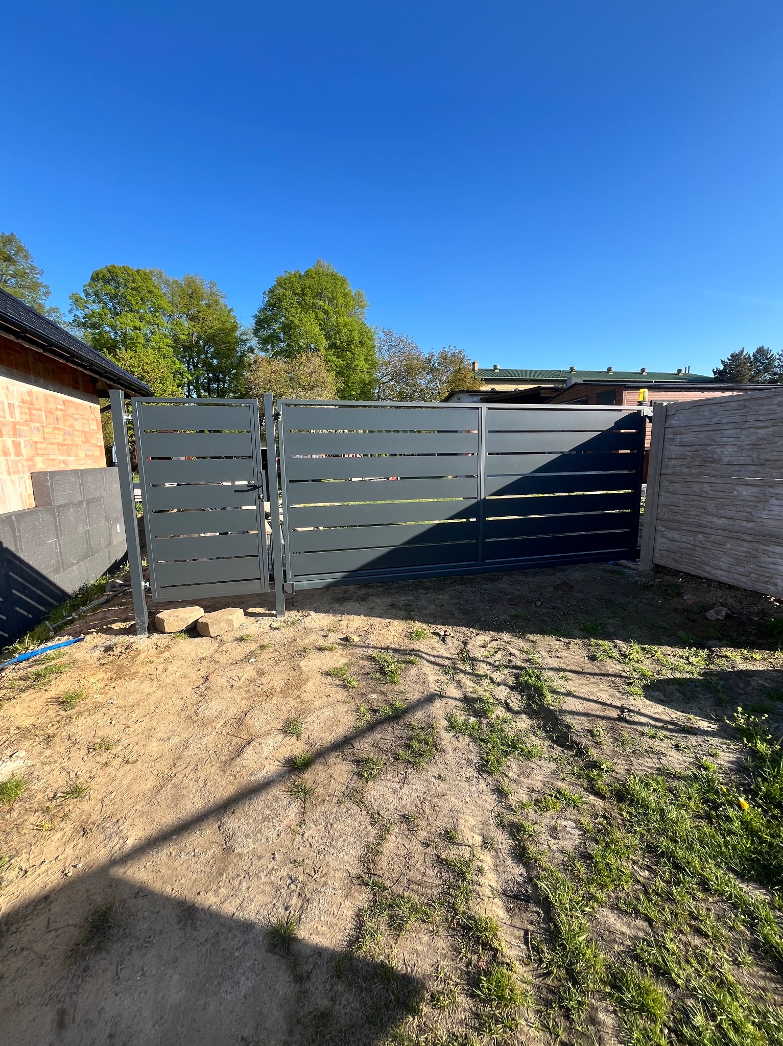 Nowoczesna, pozioma brama wjazdowa z furtką, w odcieniach szarości, na tle błękitnego nieba i zieleni. Widoczne elementy montażowe i cień na ziemi.