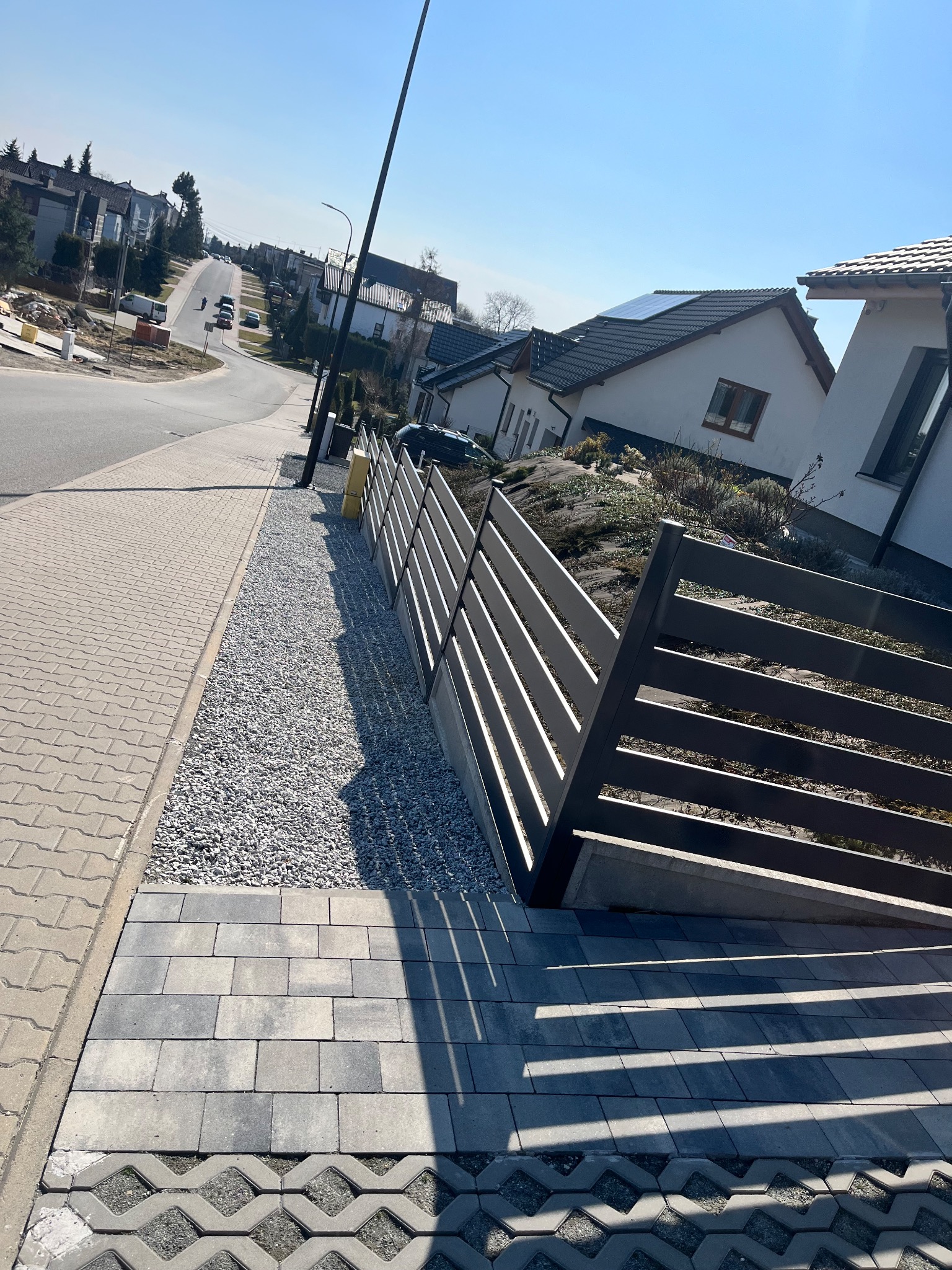 Nowoczesne, poziome ogrodzenie panelowe w kolorze grafitowym, oddzielające posesję od chodnika i ulicy. Widoczne kostki brukowe i kamień ozdobny. Ujęcie z dołu, w słoneczny dzień.