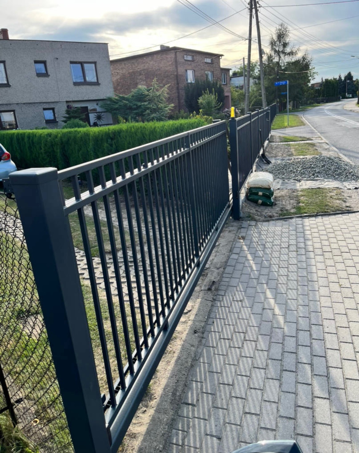Nowoczesna, przesuwna brama wjazdowa z metalowymi sztachetami w kolorze grafitowym, zamontowana przy posesji z kostką brukową i zielenią. Widok z perspektywy chodnika.